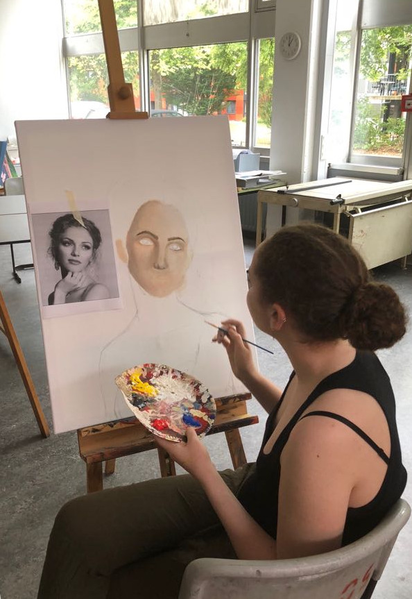 Auf dem Foto ist eine sitzende junge Frau zu sehen, die auf einer Leinwand ein Bild einer Frau malt.