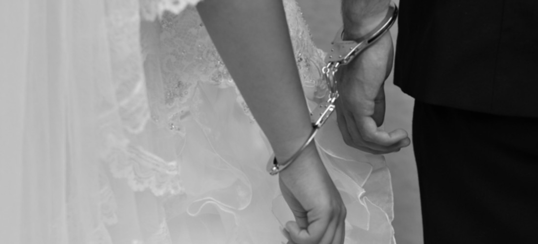 Das schwarzweiße Foto zeigt einen Ausschnitt von zwei Menschen, die mit Handschellen verbunden ist. Es handelt sich um eine Frau und einen Mann. Die Frau hat ein weißes Brautkleid an.