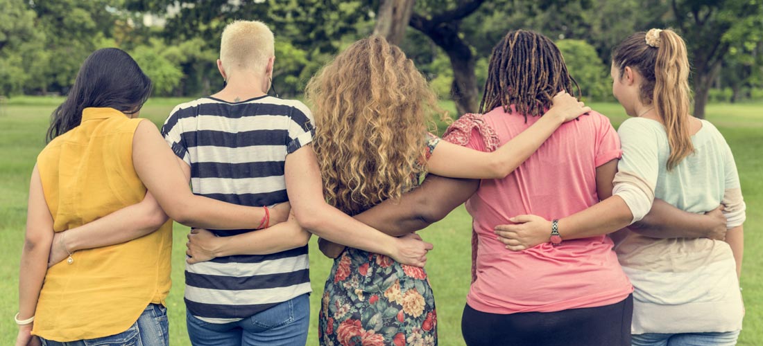 Das Foto zeigt fünf sehr unterschiedlich aussehende Frauen, die mit dem Rücken zur Kamera stehen. Sie haben sich gegenseitig die Arme um die Taille gelegt.