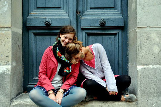 Das Foto zeigt zwei Mädchen, die im Schneidersitz vor einer Tür sitzen. Das Mädchen rechts lehnt schief an dem anderen Mädchen, ihr Kopf auf deren Schulter.