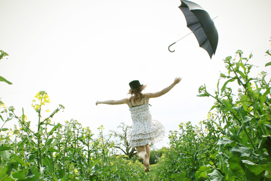 Das Foto zeigt eine junge Frau in einem weißen Kleid, die über eine Wiese läuft und einen geöffneten schwarzen Schirm von sich geworfen hat.