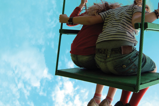 Das Foto zeigt zwei Mädchen, von hinten fotografiert, auf einer Schaukel vor strahlendblauem Himmel. 