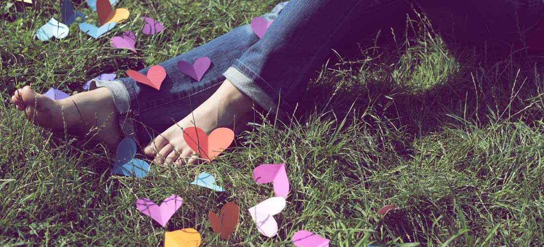 Das Foto zeigt die Beine von einem Mädchen auf einer Wiese. Über ihre Füße verstreut liegt großes, buntes Herzkonfetti.